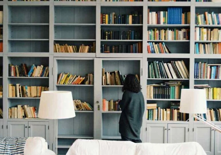 woman looking at all the stuff on her big bookshelf thinking about downsizing